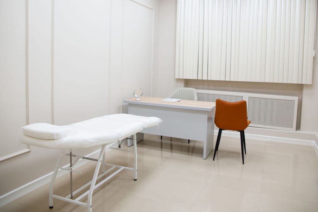 Ljust behandlingsrum på klinik med undersökningssäng och skrivbord för konsultation.
